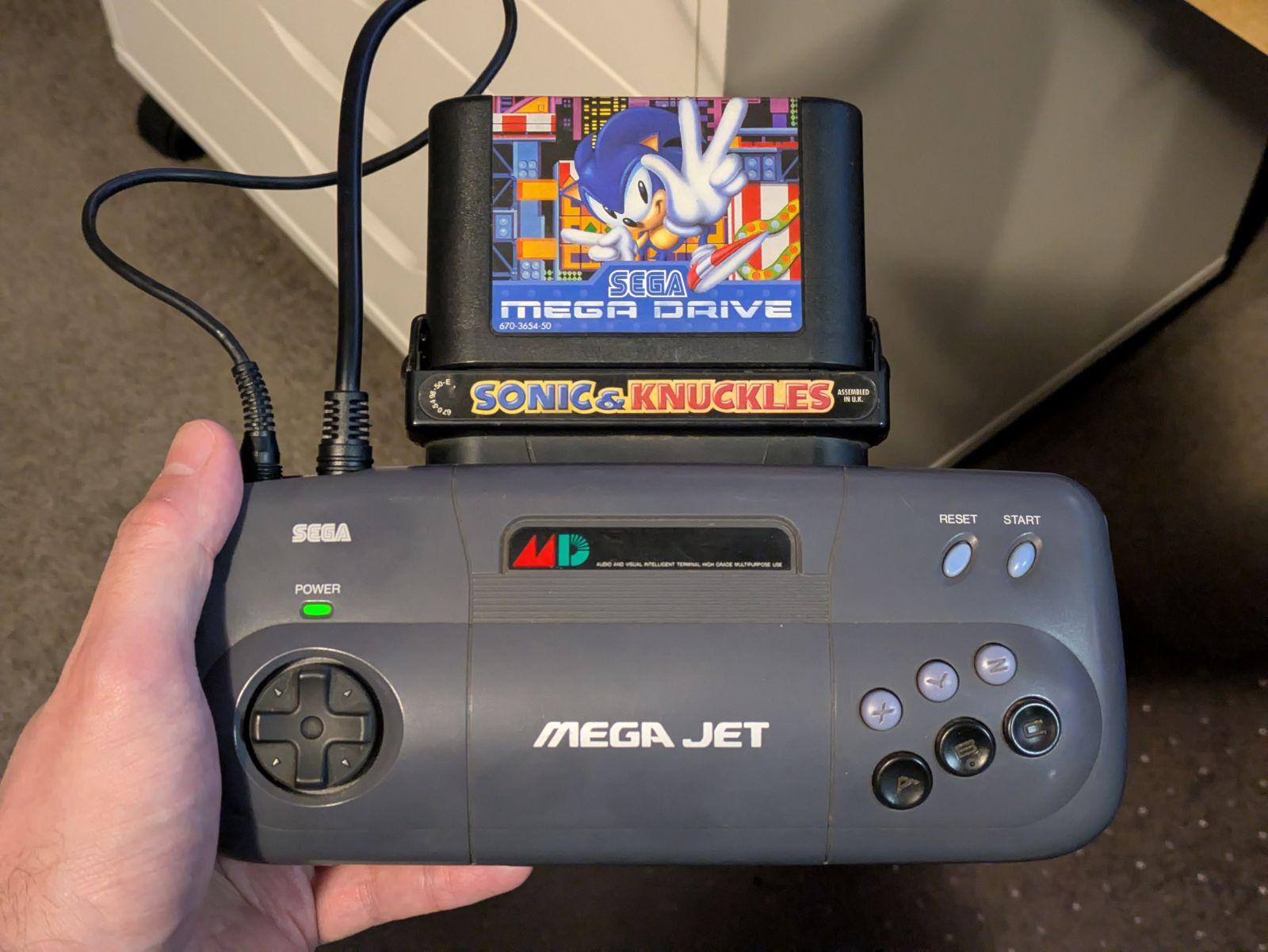 A photo of a Sega Mega Jet console. It's a large handheld device with a D-pad on the left, six action buttons on the right, and two cables and a large game cartridge sticking out of the top.