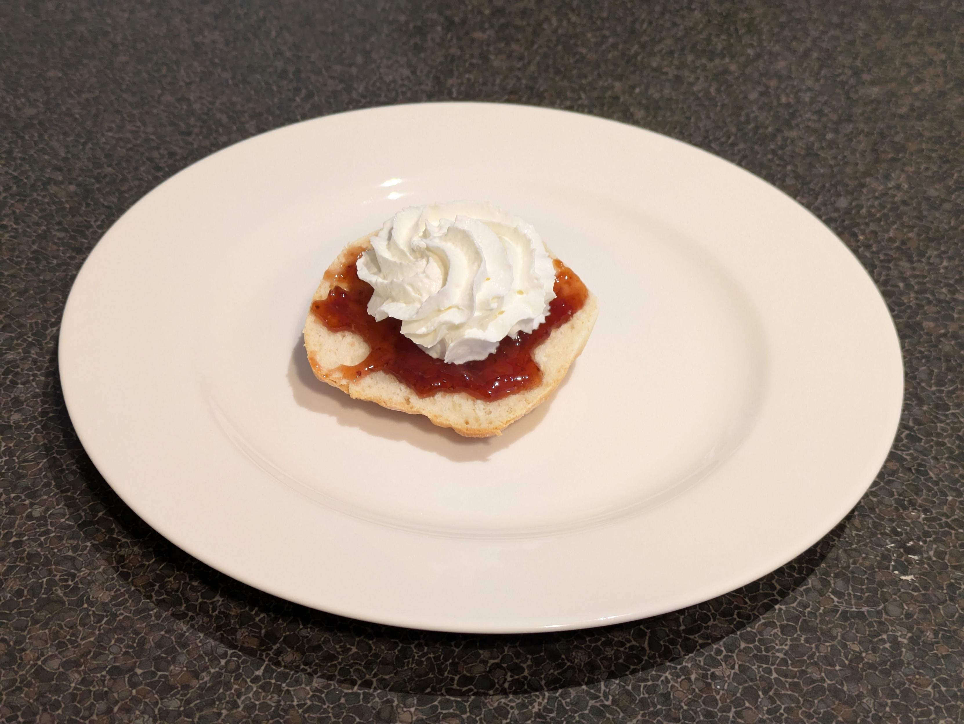 A photo of half a baked scone on a plate. It's been topped prettily with strawberry jam and whipped cream.